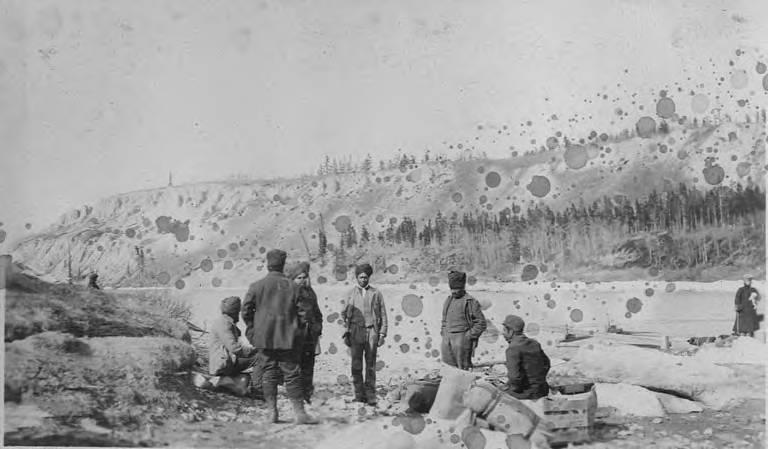 April 1906, Sikh men in turbans sitting and standing on rocky bank with bags and supplies. Whitehorse, Yukon.